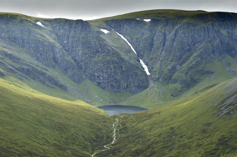 Rock cliffs leading down to a loch, with some snow in the crevices.