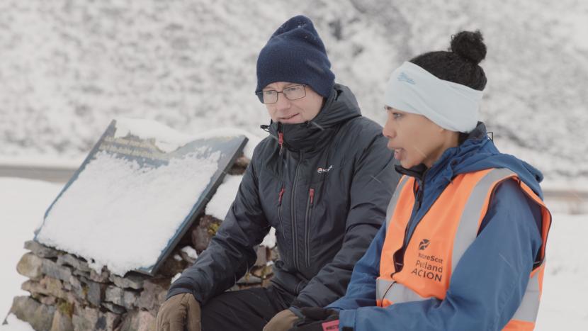 Two people talking in the snow, wearing winter clothing and a high visibility vest.