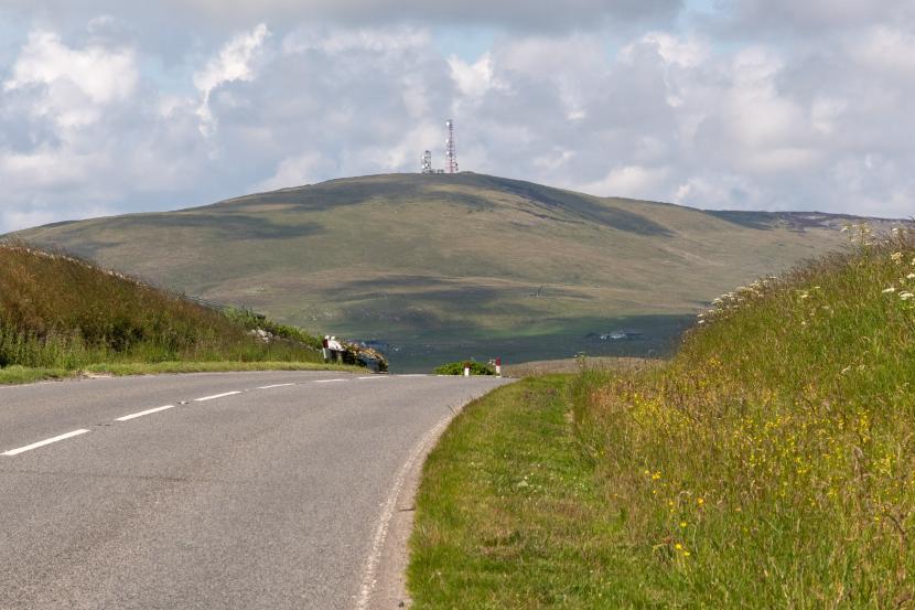 Strech of road over the top of a hill, with wildflowers on the verge and a hill in the distance.