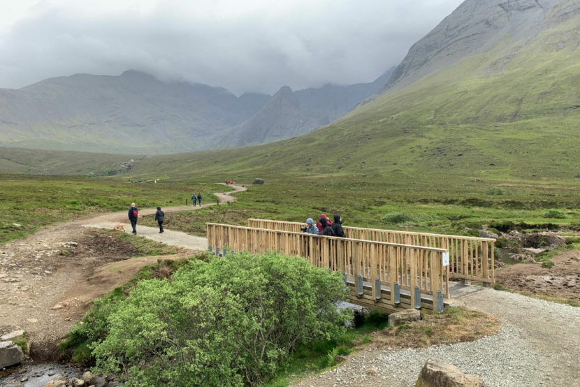 Walkers using a gravel path and crossing a bridge, with mountains in the distance.