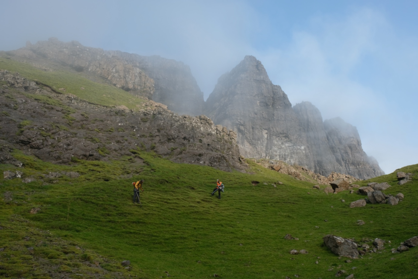 2 workers in high vis jackets, working on a hill side with steep cliffs in the background.