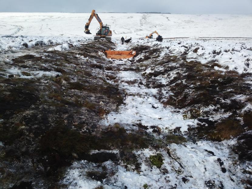 View looking down a wide ditch in snowy conditions. In the background two machine operators are reprofiling the bare steep edges of the ditch. Halfway along the ditch a timer dam has been installed to slow the flow of water and trap sediments.