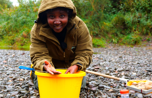 A person on a pebble beach inspecting items in a bucket - there is a fishing net beside them.