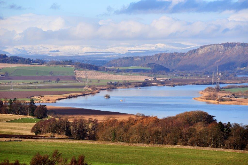View over a river with snowy mountains in the distance and farmland in the foreground.