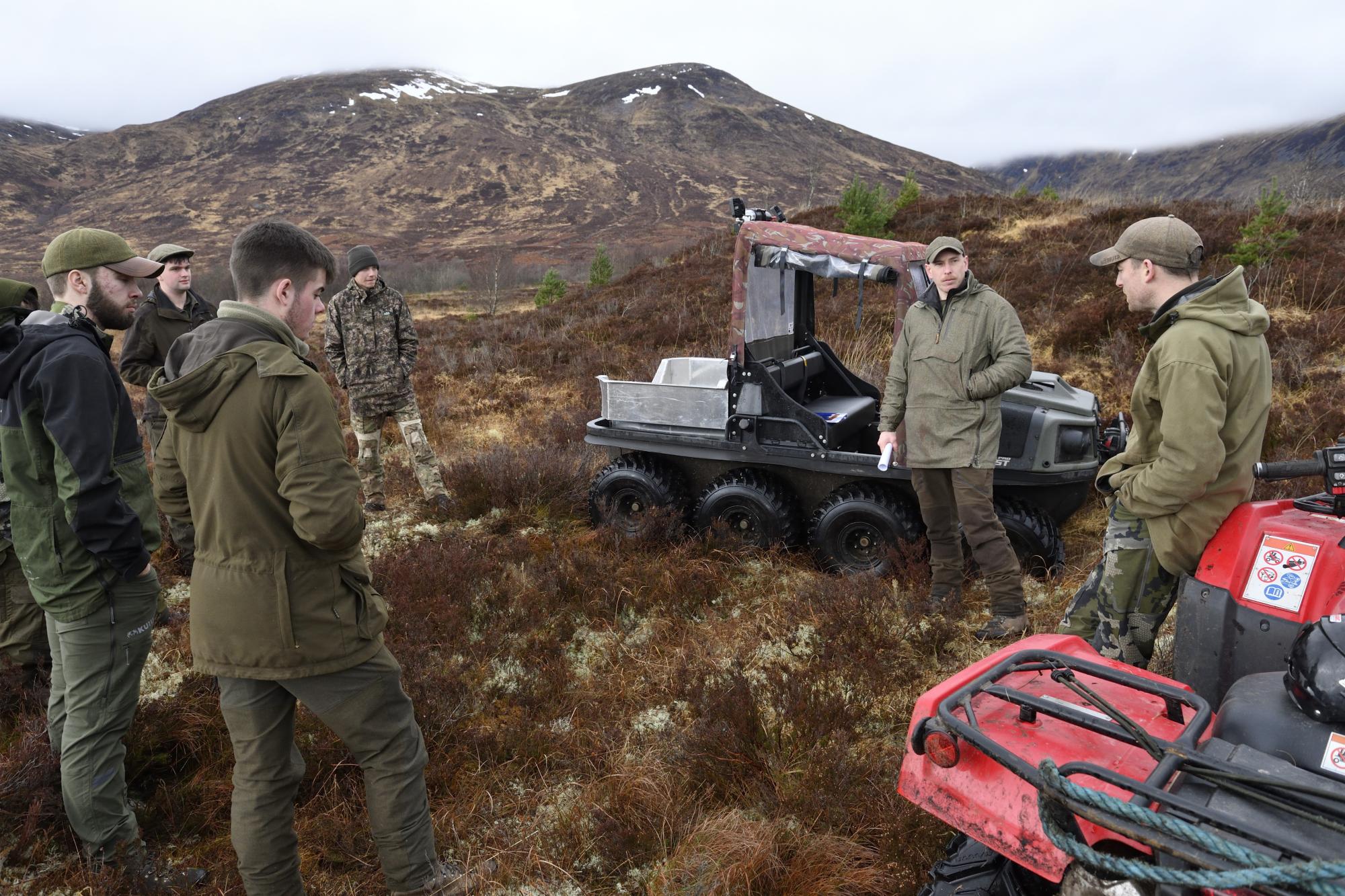 6 men in green clothing standing on a hillside, with 2 off road buggies. Snow covered mountains in the background.
